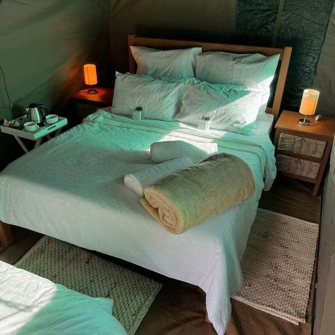 A double bed with white linen, folded towels and warm bedside lamps inside a tent
